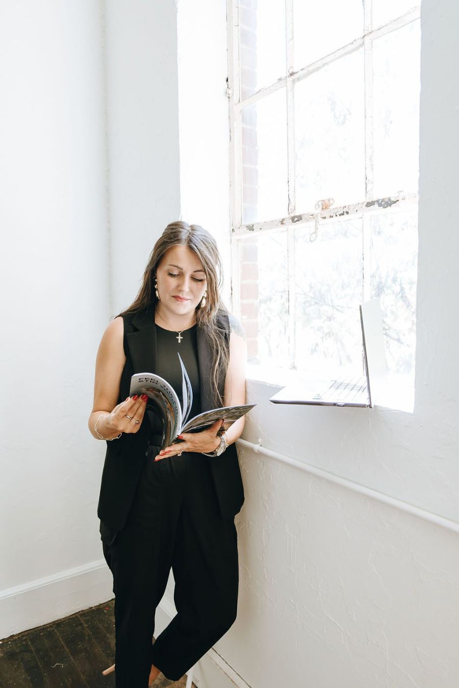 Emily reading a magazine by a tall sunlit window, laptop on the sill