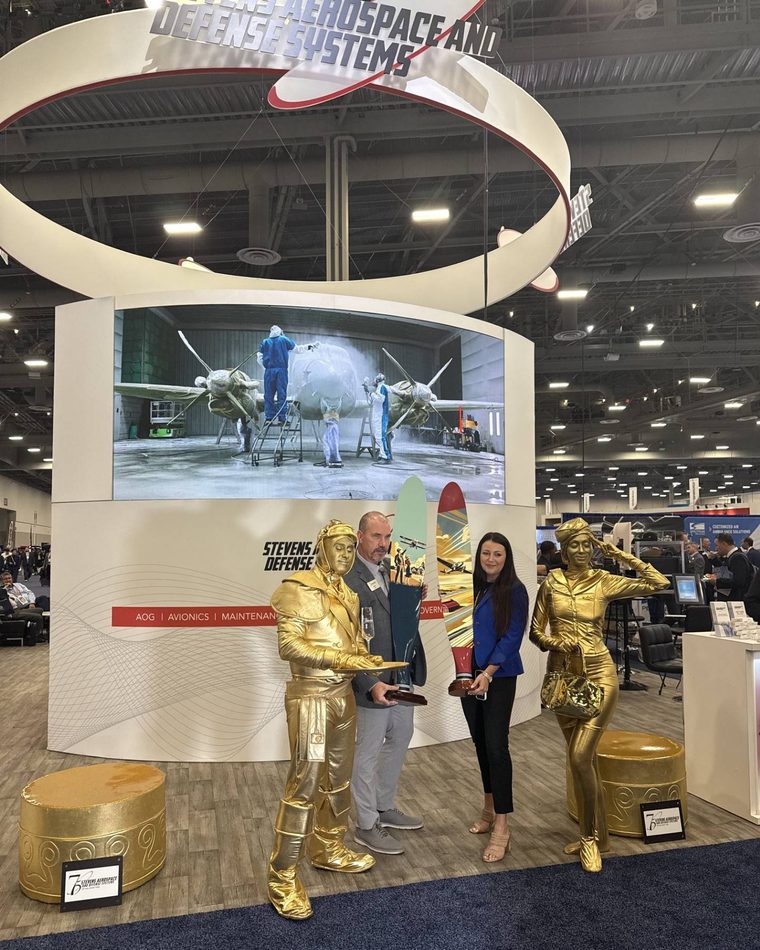The Stevens Aerospace and Defense Systems booth at NBAA-BACE, with its hanging sign, video wall, and gold living statues