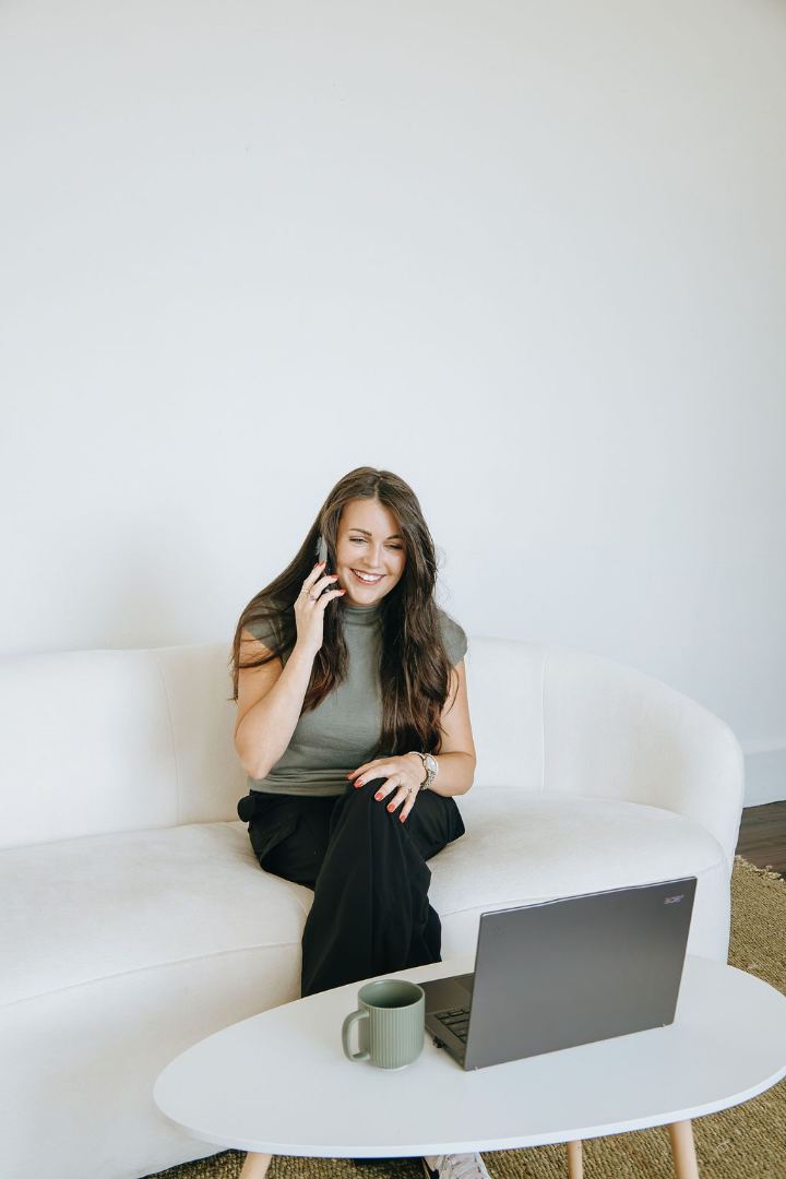 Emily smiling on a phone call, seated on a sofa with a laptop and coffee